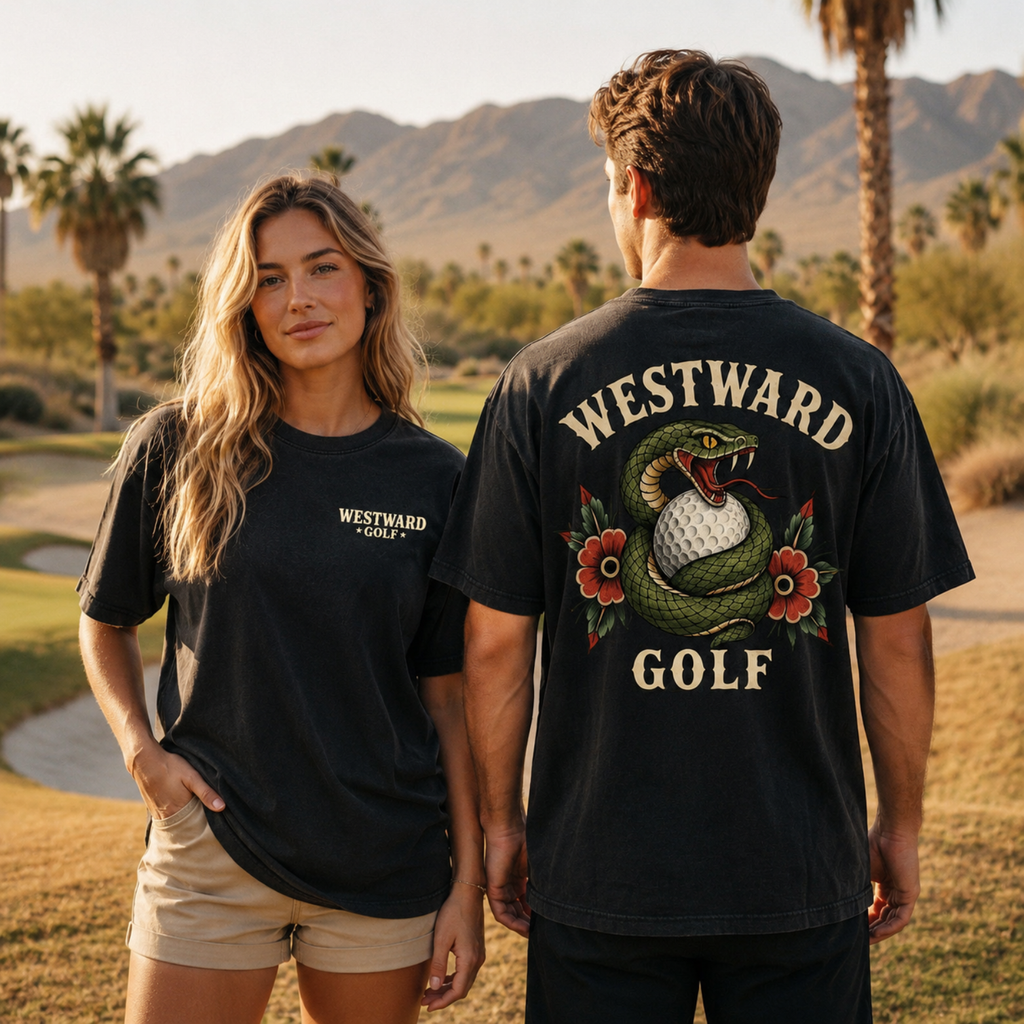 Male and female models wearing black Westward Golf t-shirts on a desert golf course, with the woman facing forward to show the small chest logo and the man facing away to showcase the large snake and golf ball back graphic.