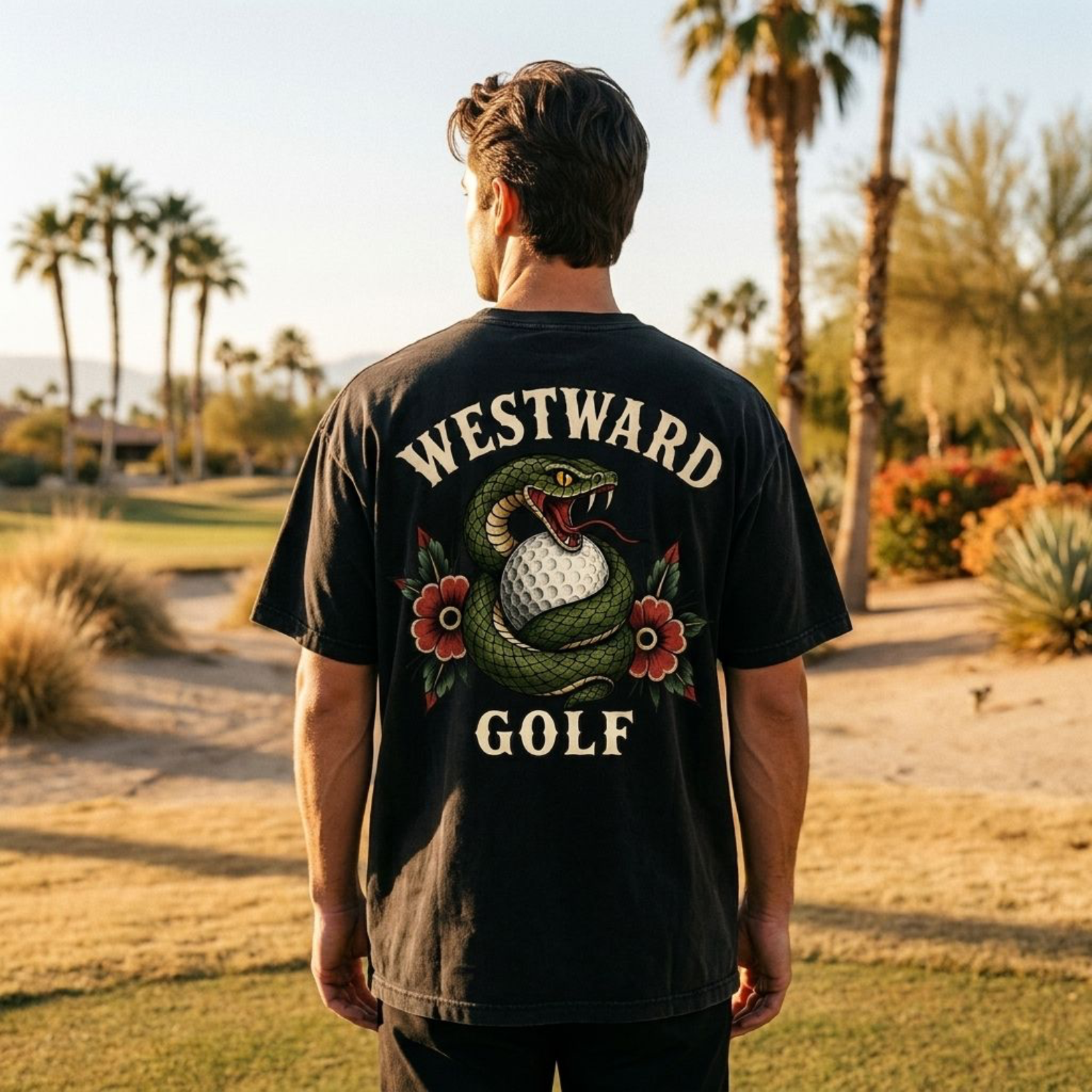 Male model wearing a black Westward Golf t-shirt facing away on a desert golf course beside a golf cart, showcasing the large snake and golf ball back graphic with cream lettering, red flowers, and warm sunset lighting.