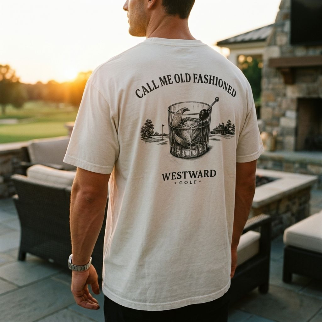 Male model wearing an ivory Westward Golf t-shirt from the back, featuring a “Call Me Old Fashioned” old fashioned cocktail graphic and Westward Golf text, standing on an upscale golf club patio at sunset.
