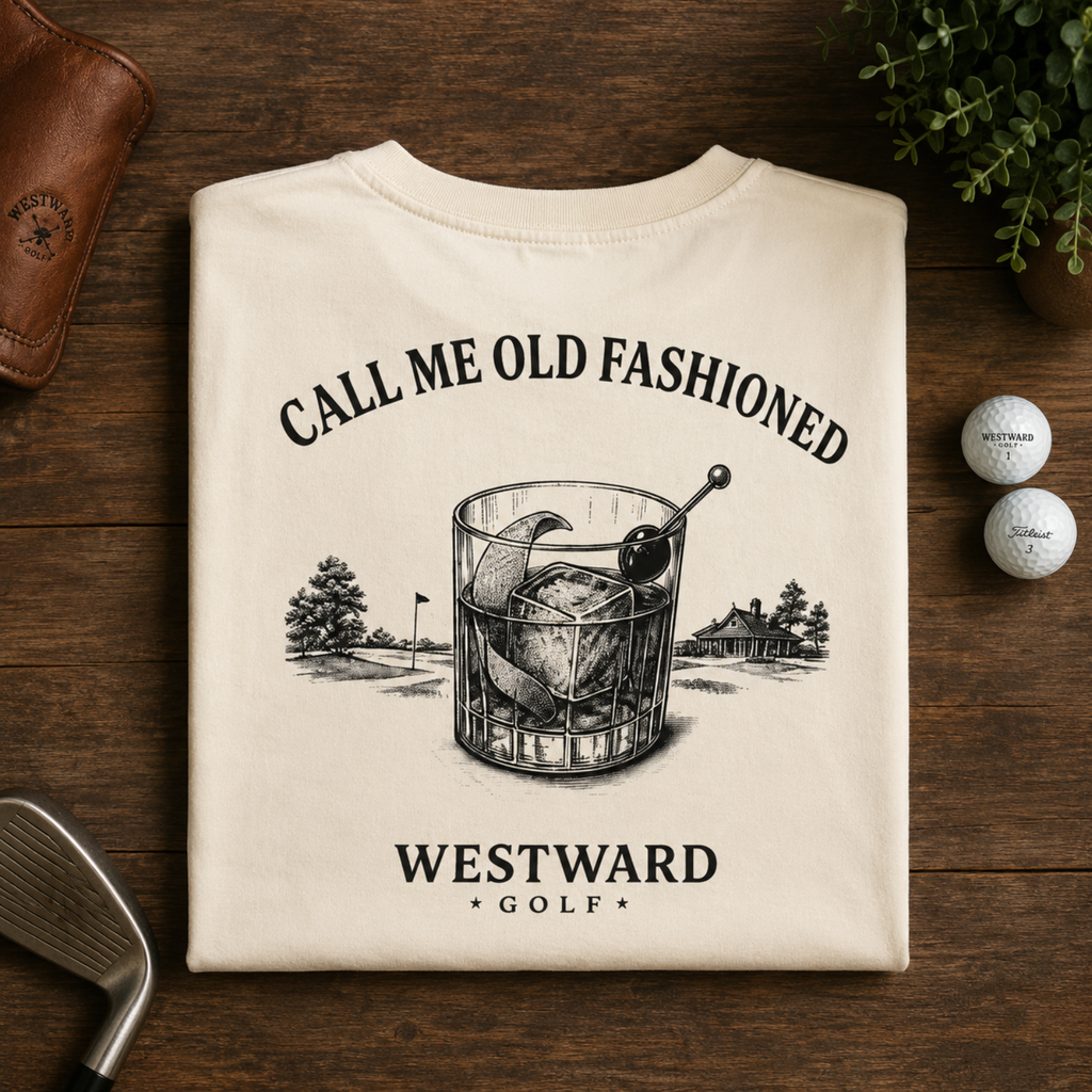 Folded ivory Westward Golf t-shirt displayed on a dark wood flat lay, featuring a large “Call Me Old Fashioned” back graphic with an old fashioned cocktail glass, golf course details, and Westward Golf text, styled with golf balls, a golf club, greenery, and a leather accessory.