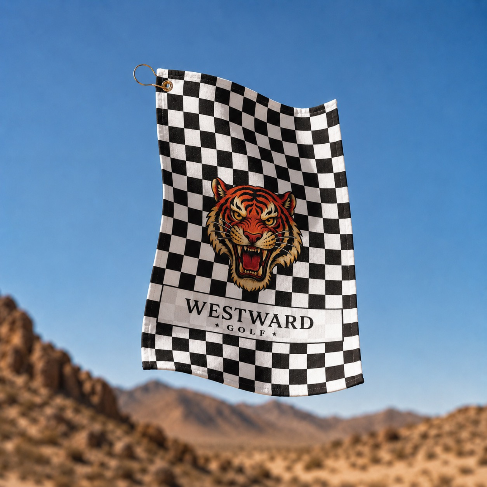 Black and white checkered Westward Golf towel with bold vintage roaring tiger logo, premium golf bag towel flying outdoors with desert mountain background, checkerboard golf towel accessory with gold hanging ring and brass grommet for cleaning golf clubs, golf balls, grips, hands, and golf gear on the course.