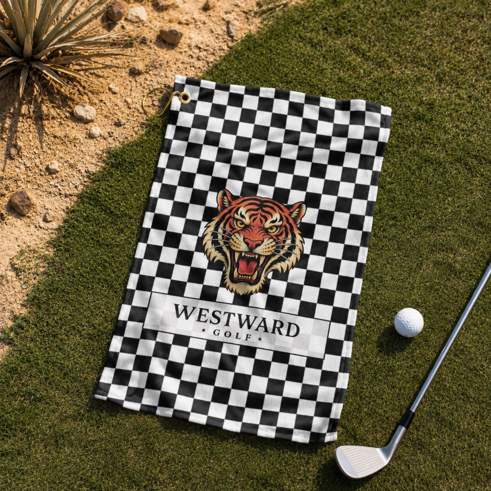 Black and white checkered Westward Golf towel laid on green golf course grass with vintage roaring tiger logo, premium checkerboard golf towel with gold hanging ring and brass grommet, stylish golf accessory beside golf ball and club for cleaning clubs, golf balls, grips, hands, and gear during every round.