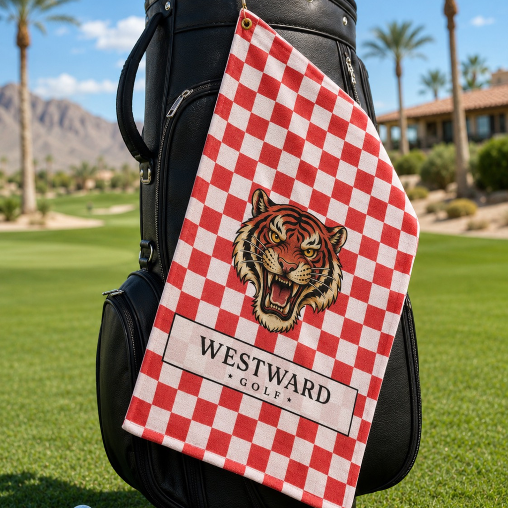 Red and white checkered Westward Golf towel hanging from a black golf bag on a sunny golf course, featuring bold vintage roaring tiger logo, premium checkerboard golf towel with brass grommet and gold hanging ring, stylish golf bag accessory for cleaning golf clubs, golf balls, grips, hands, and gear during every round.