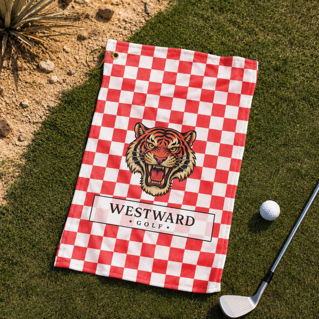 Red and white checkered Westward Golf towel laid on green golf course grass with vintage roaring tiger logo, premium checkerboard golf towel with gold hanging ring and brass grommet, bold golf accessory beside golf ball and club for cleaning golf clubs, golf balls, grips, hands, and gear during every round.