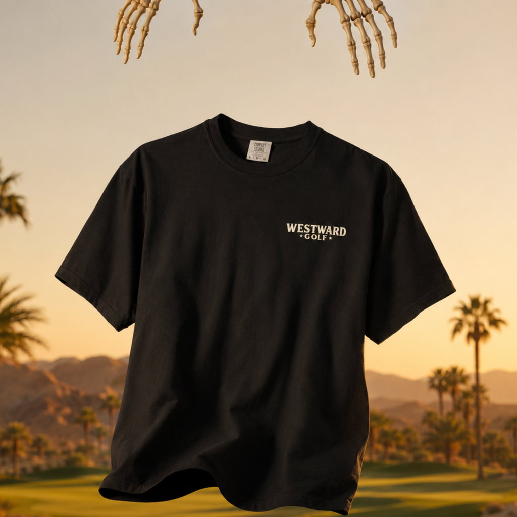 Black Westward Golf t-shirt floating over sunset golf course with small front chest logo, skeleton hands above, palm trees, and desert mountain background
