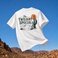 White Westward Golf “The Twilight Special” graphic T-shirt mockup featuring vintage desert golf artwork with green lettering, cactus, orange sunset, mountains, and golf bag, shown floating against a blue sky and desert mountain background.
