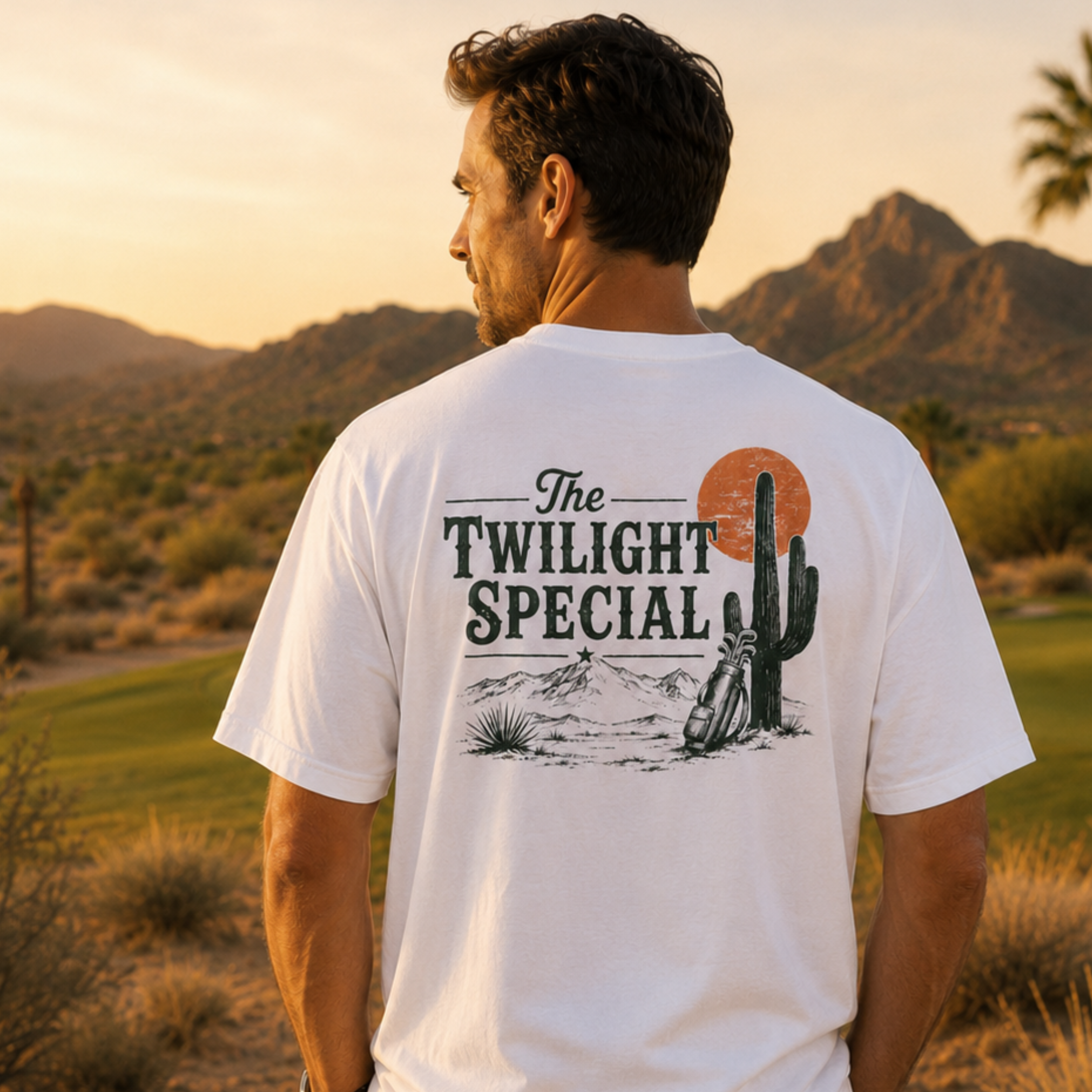 Male model wearing white Westward Golf “The Twilight Special” graphic T-shirt on a desert golf course at sunset, showing vintage back print with cactus, orange sun, mountains, and golf bag design.