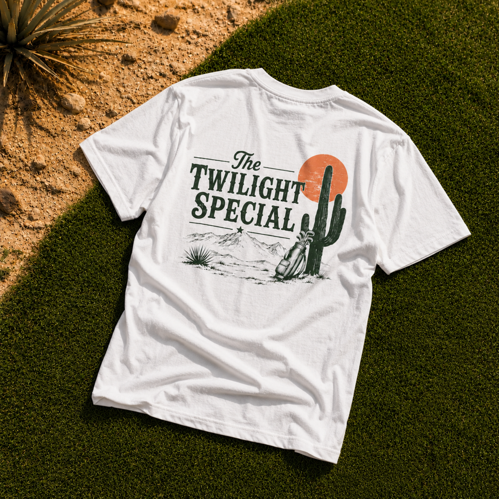 Flat lay white Westward Golf “The Twilight Special” graphic T-shirt on green golf course grass with desert sand, rocks, and cactus plant, featuring vintage back print with green lettering, orange sunset, mountains, cactus, and golf bag design.